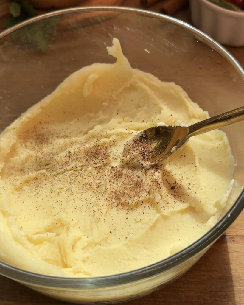 Smooth mashed potatoes in a large glass bowl, topped with a sprinkle of spices, ready for serving.