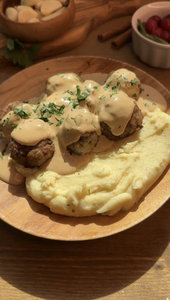 A wooden plate featuring juicy homemade meatballs covered in creamy gravy, served alongside buttery mashed potatoes, garnished with freshly chopped parsley for a flavorful and visually appealing dish.