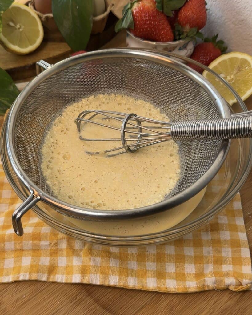 Straining crepe batter through a sieve to remove lumps, ensuring smooth and soft crepes for strawberries and cream crepe rolls