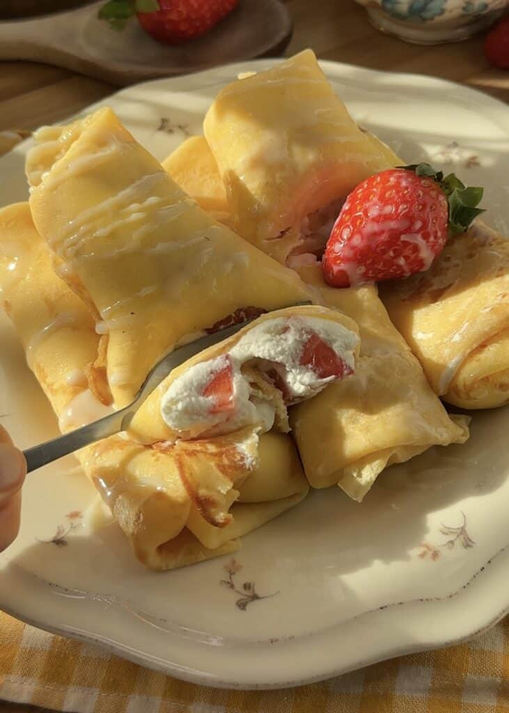 A fork cutting into a strawberries and cream crepe roll, revealing the creamy whipped filling and fresh strawberry slices inside.