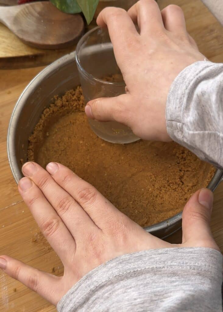 Pressing down the crust for a no-bake pistachio cheesecake with a glass in a cheesecake pan, preparing it to be chilled in the freezer while the filling is made