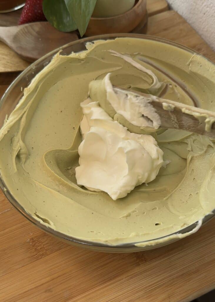 A bowl with a creamy mixture of cream cheese and pistachio spread, topped with heavy cream ready to be incorporated into the filling for a smooth no-bake pistachio cheesecake.