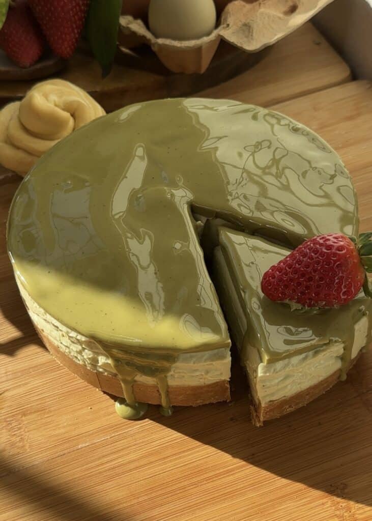 A no-bake pistachio cheesecake with a creamy filling on a wooden board, topped with pistachio spread and a fresh strawberry, with a cut slice revealing its smooth texture.