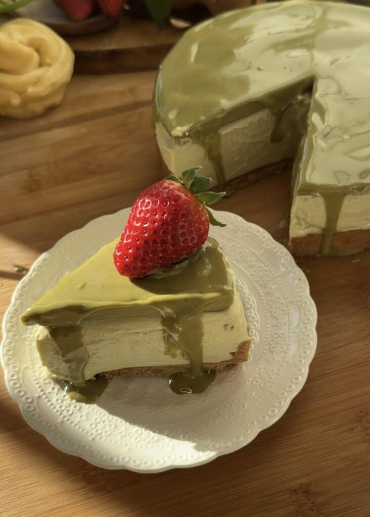 Sliced no bake pistachio cheesecake on a plate with three layers: crust, creamy filling, pistachio topping, and fresh strawberry garnish. Full cheesecake in background