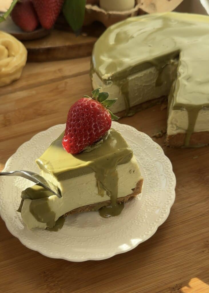 A slice of no-bake pistachio cheesecake with three layers: a buttery crust, a creamy pistachio cheesecake filling, and a pistachio spread topping. A spoon sinks into the smooth and creamy texture. Garnished with a fresh strawberry on a white plate.