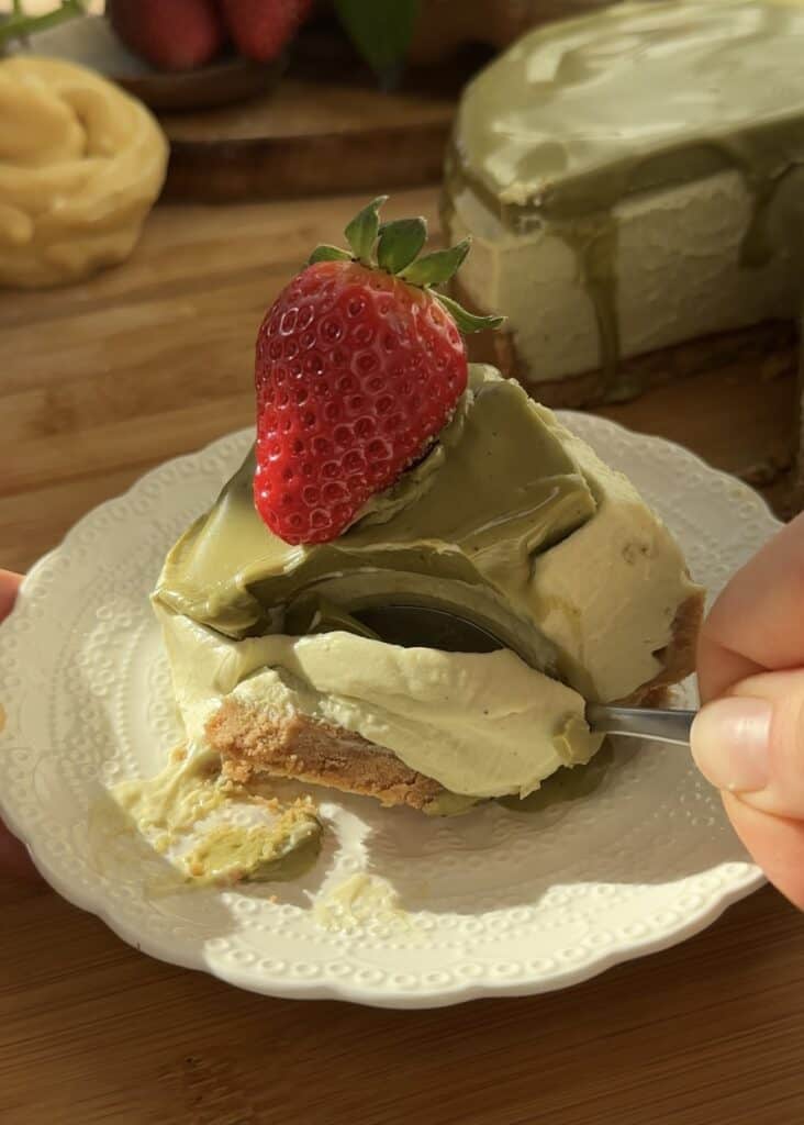 A slice of no-bake pistachio cheesecake with three layers: a buttery crust, a creamy pistachio cheesecake filling, and a pistachio spread topping. A spoon sinks into the smooth and creamy texture. Garnished with a fresh strawberry on a white plate.