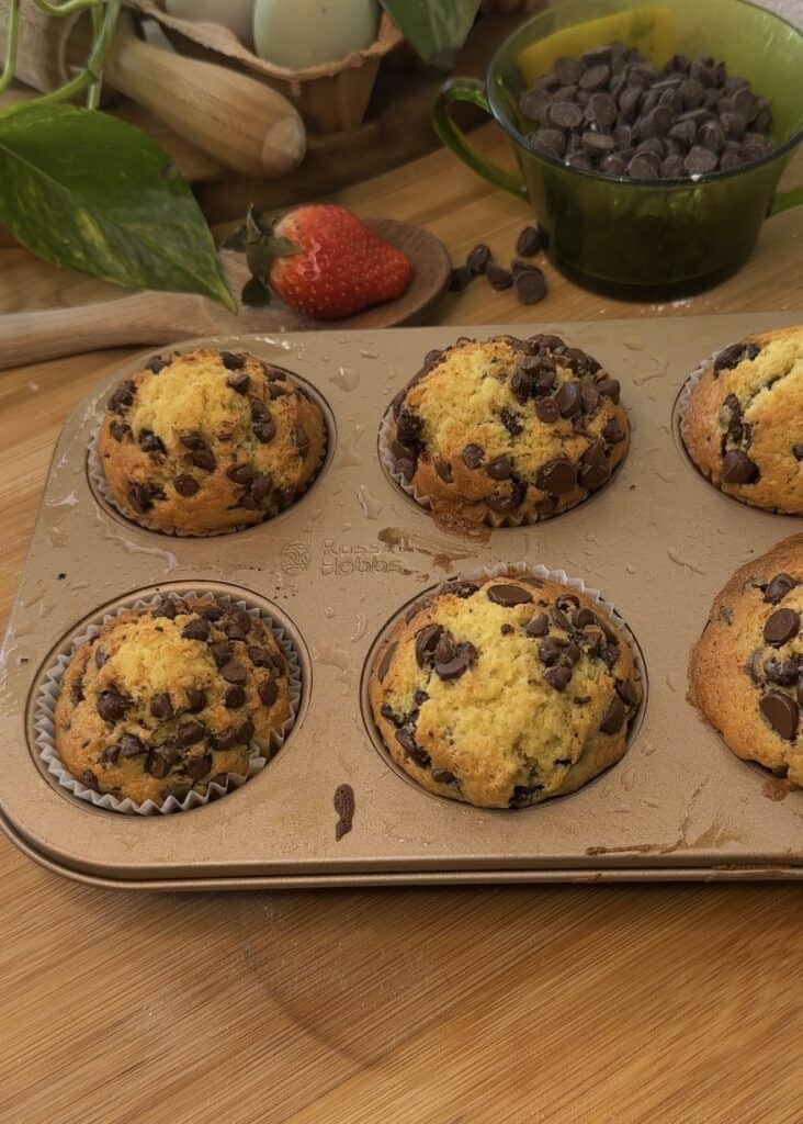 Freshly baked muffins in a pan with chocolate chips, ready for Nutella filling using a piping bag.