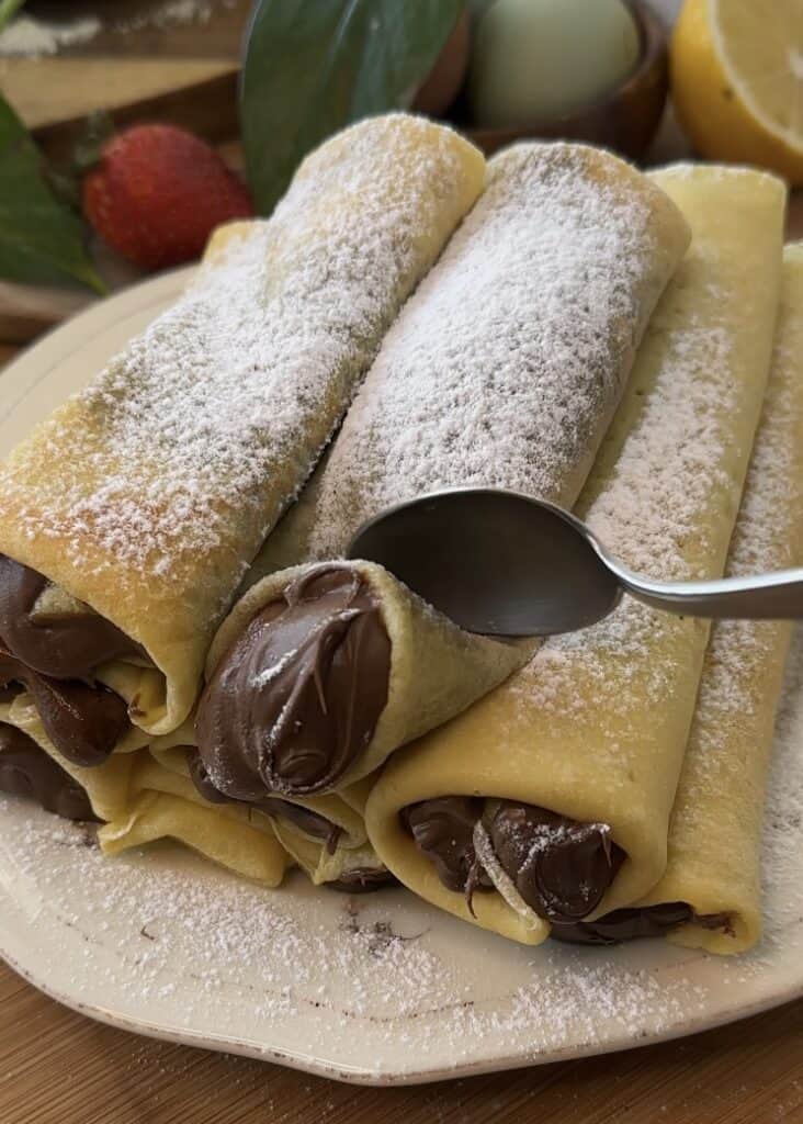 Stack of Nutella crepe rolls on a plate with Nutella oozing out and powdered sugar on top, ready to serve