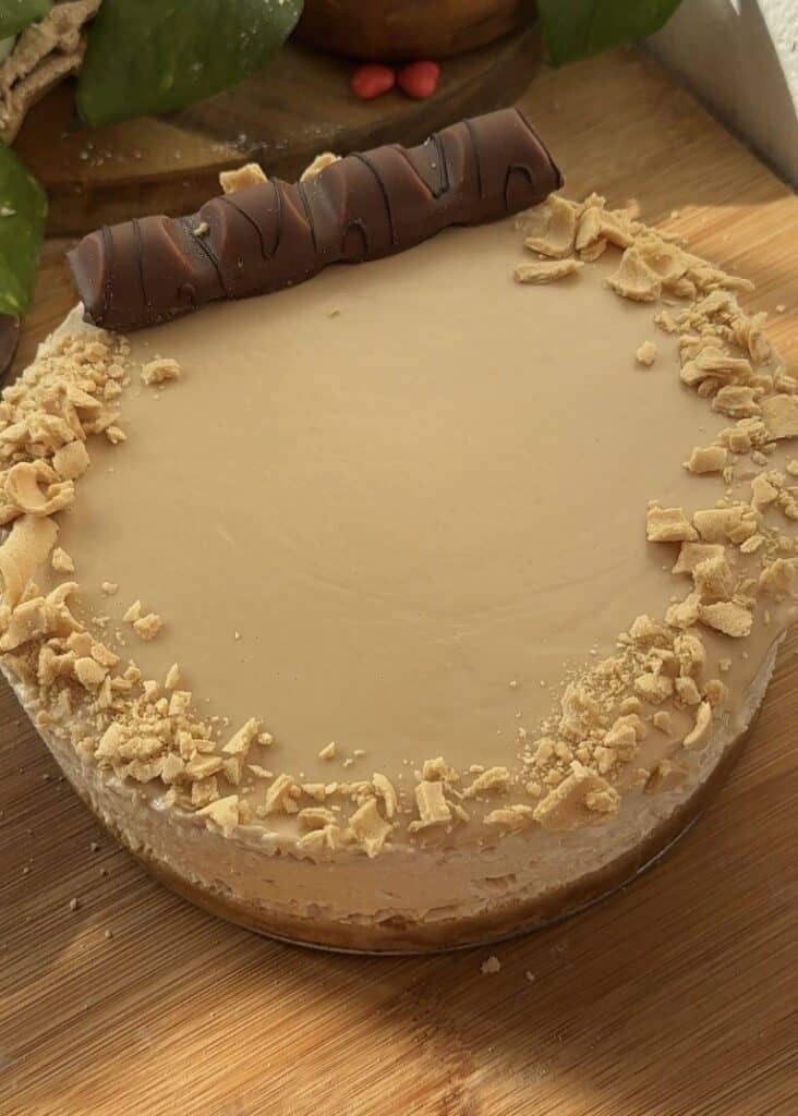 No-bake Kinder Bueno cheesecake with a whole Kinder Bueno bar placed on one side, showcasing its crispy wafer and creamy hazelnut filling. Simple presentation focusing on the bar as the centerpiece.