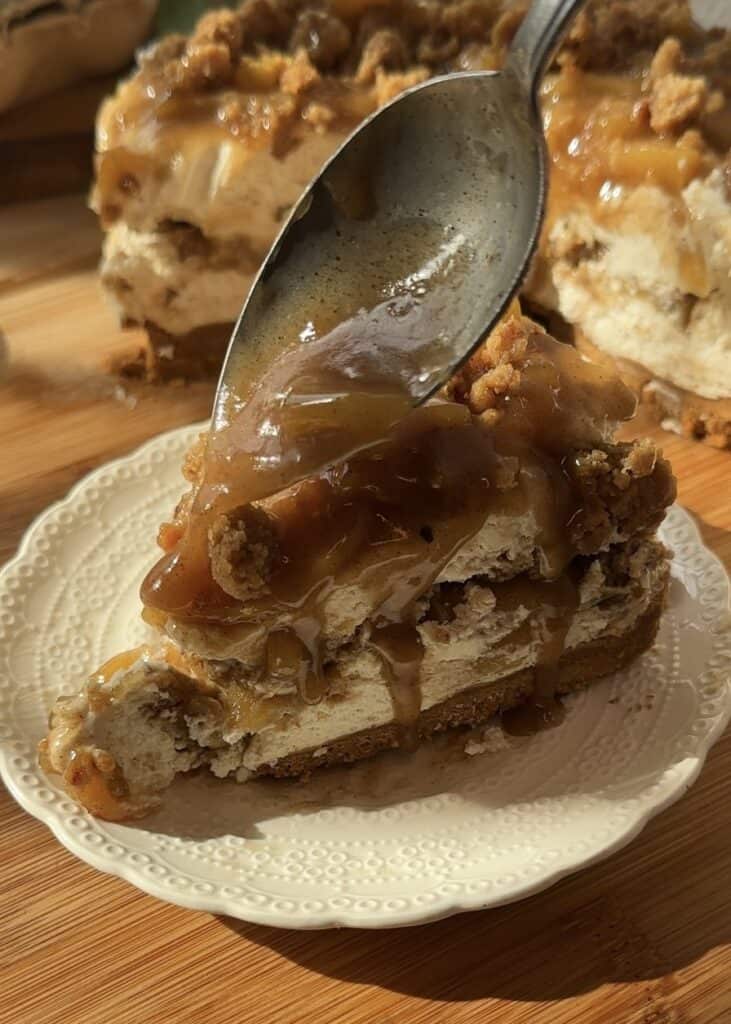 A spoon scooping the first bite of a slice of no-bake apple pie cheesecake with crumble. The creamy cheesecake has a crunchy crumble base, a smooth cheesecake layer, spiced saucy apples in the middle, and a topping of more saucy apples and crumble.