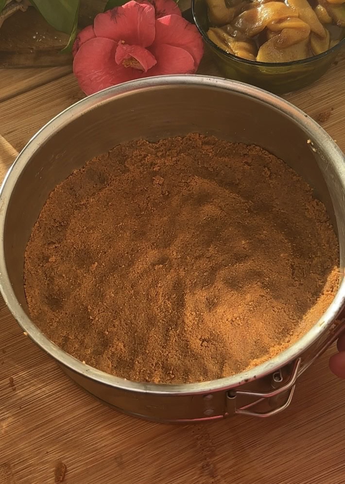 Pressing crushed cookies mixed with butter into a greased baking mold with baking paper to form the crust for no-bake apple pie cheesecake with crumble.