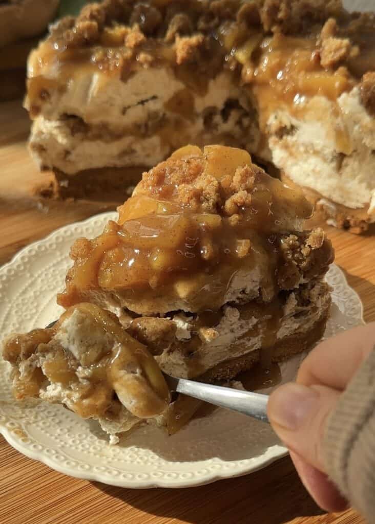 A spoon scooping the first bite of a slice of no-bake apple pie cheesecake with crumble. The creamy cheesecake has a crunchy crumble base, a smooth cheesecake layer, spiced saucy apples in the middle, and a topping of more saucy apples and crumble.