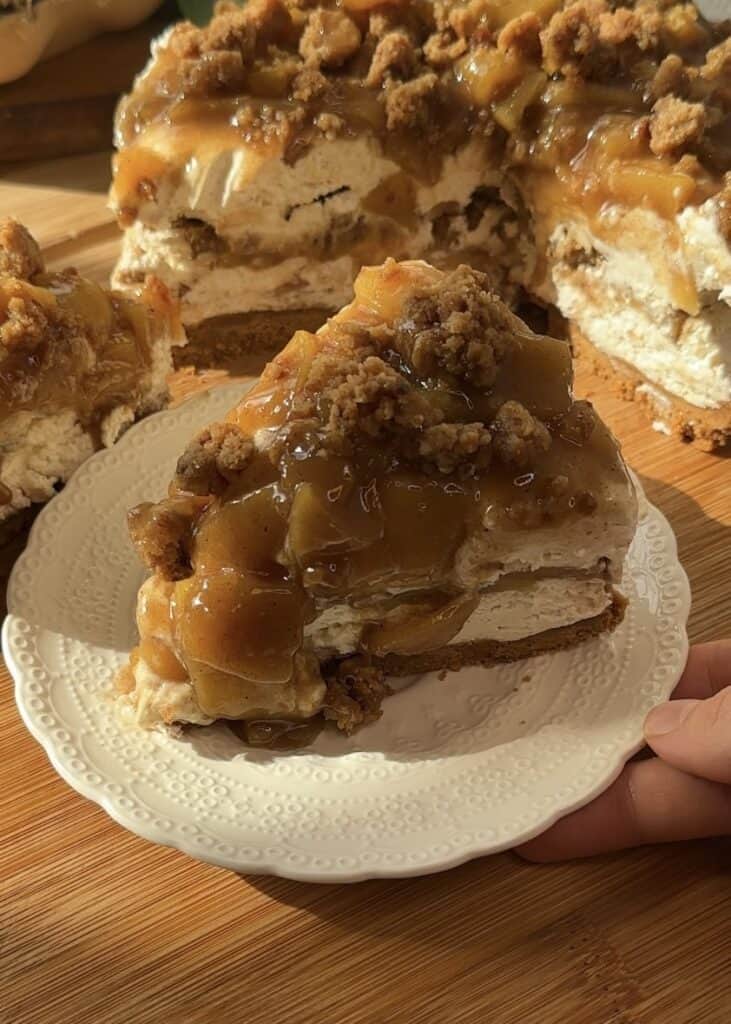 A slice of no-bake apple pie cheesecake with crumble, featuring a crunchy crumble base, smooth cheesecake filling, spiced saucy apples in the middle, and topped with more saucy apples and crumble.