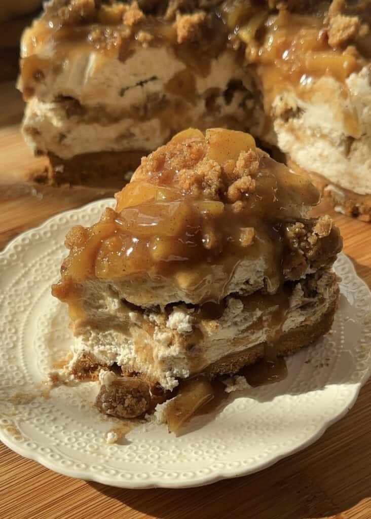 A spoon scooping the first bite of a slice of no-bake apple pie cheesecake with crumble. The creamy cheesecake has a crunchy crumble base, a smooth cheesecake layer, spiced saucy apples in the middle, and a topping of more saucy apples and crumble.