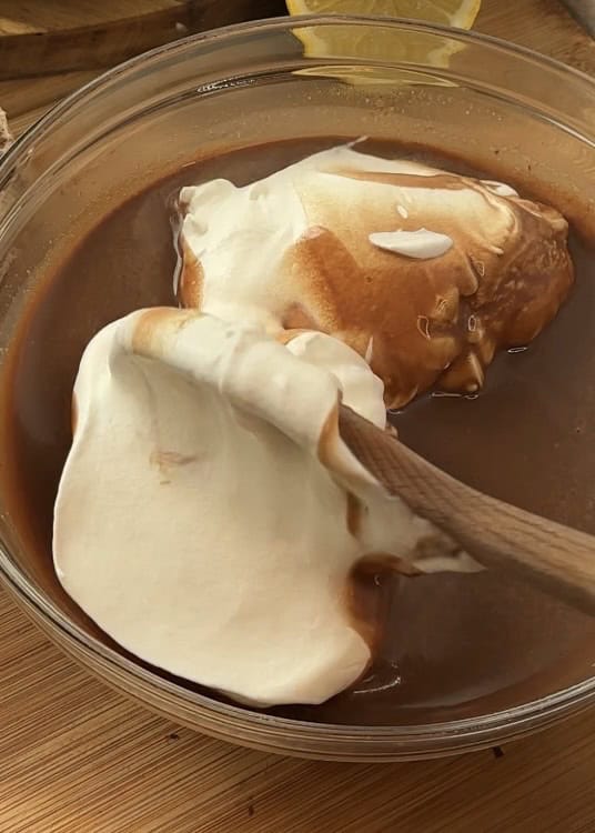 Mixing milk chocolate cream with whipping cream using a wooden spoon in a glass bowl to create a smooth, rich milk chocolate mousse for the cake.