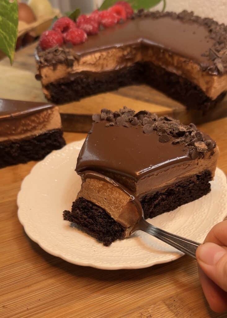 A spoon scooping the second bite of a slice of milk chocolate mousse cake, with a chocolate cake base, milk chocolate mousse filling, ganache topping, chocolate chips, fresh raspberries, and mint leaves.