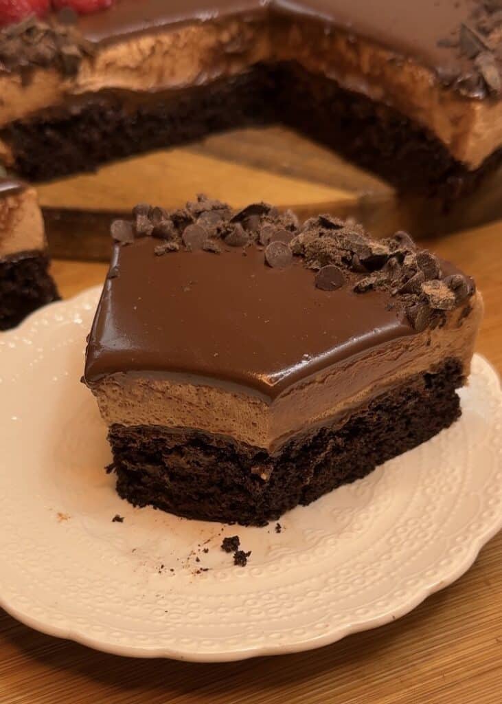 A slice of milk chocolate mousse cake with a chocolate cake base, milk chocolate mousse filling, topped with ganache, chocolate chips, fresh raspberries, and mint leaves for decoration.