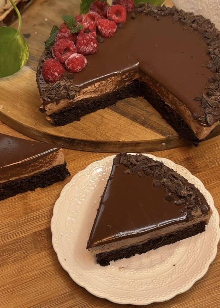 A slice of milk chocolate mousse cake with a chocolate cake base, milk chocolate mousse filling, topped with ganache, chocolate chips, fresh raspberries, and mint leaves for decoration.