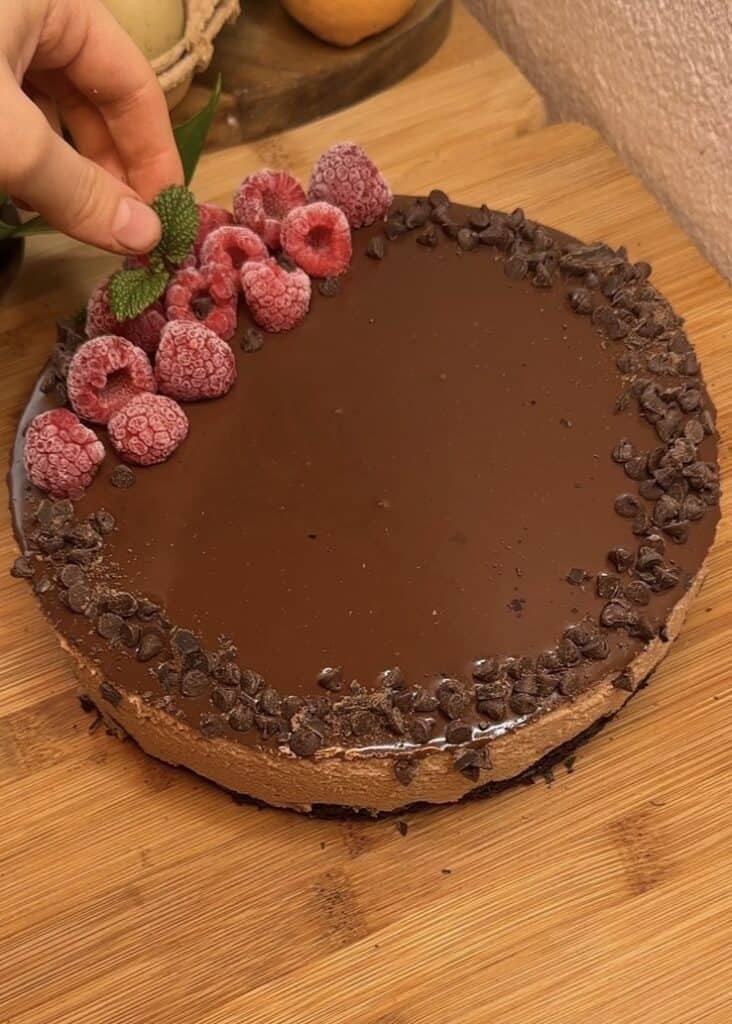 A milk chocolate mousse cake with a chocolate cake base, milk chocolate mousse filling, topped with ganache, chocolate chips, fresh raspberries, and mint leaves for decoration.
