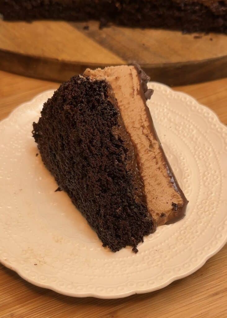 A slice of milk chocolate mousse cake with a chocolate cake base, milk chocolate mousse filling, topped with ganache, chocolate chips, fresh raspberries, and mint leaves for decoration.