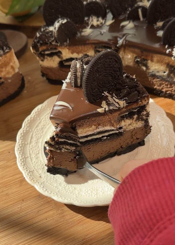 A spoon scooping the first bite of a slice of no-bake chocolate Oreo cheesecake with a crunchy Oreo crust, layers of milk chocolate and white chocolate cheesecake, Oreo pieces in the middle, and topped with smooth ganache, whole Oreos, and grated white chocolate.