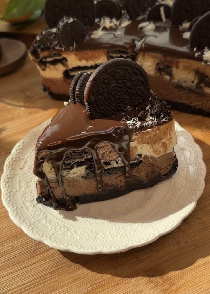 A slice of no-bake chocolate Oreo cheesecake with a crunchy Oreo crust, layers of milk chocolate and white chocolate cheesecake, Oreo pieces in the middle, and topped with smooth ganache, whole Oreos, and grated white chocolate.