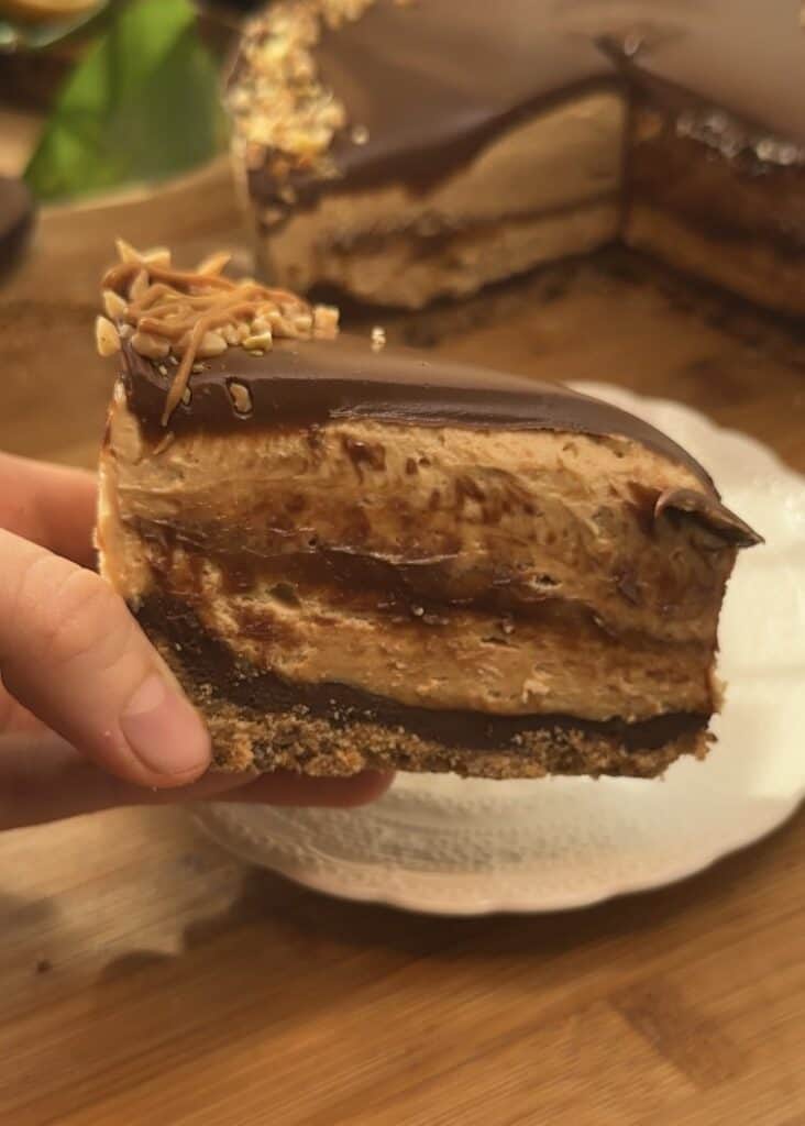 Slice of Peanut Butter Cheesecake on a plate, showing a perfect cookie crust, smooth peanut butter cheesecake, a rich peanut butter middle layer, and topped with Nutella and crushed peanuts, highlighting all the layers and creamy texture.