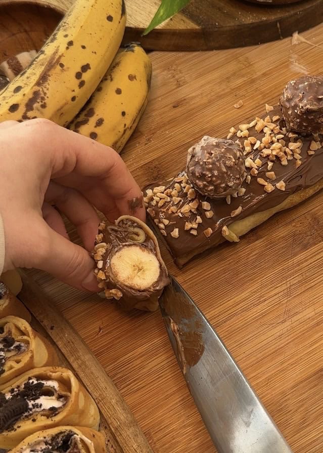 Close-up of cutting sushi crepes filled with Nutella, Ferrero Rocher, and banana slices, topped with extra Nutella drizzle and granulated almonds for decoration, ready to serve on a wooden table