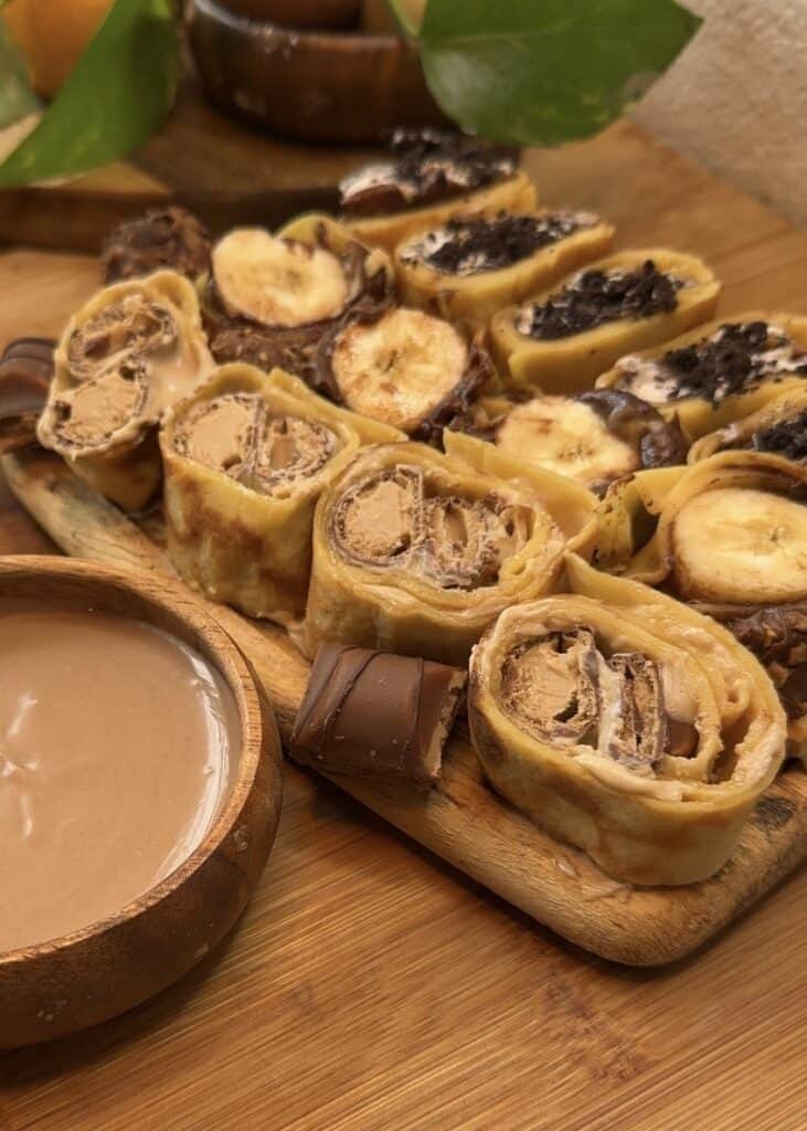 Sushi crepes on a wooden table with three varieties: Kinder Bueno spread and crushed Kinder Buenos, Nutella and banana with Ferrero Rocher, and whipped cream with chocolate and crushed Oreos, served with a bowl of Kinder Bueno spread for dipping