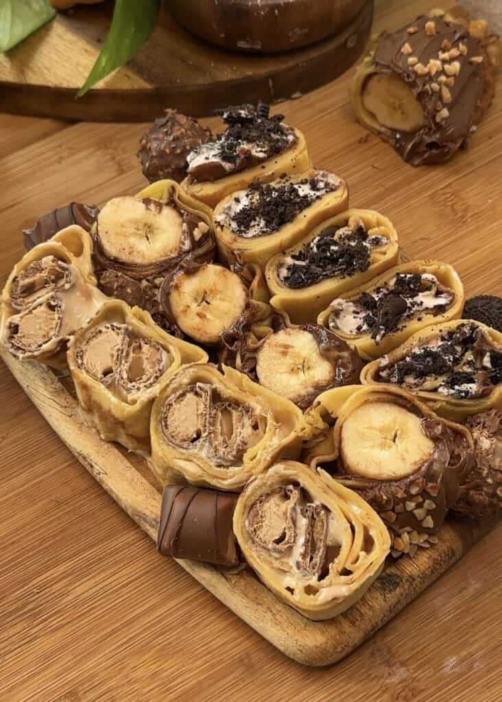 Sushi crepes on a wooden table with three varieties: Kinder Bueno spread and crushed Kinder Buenos, Nutella and banana with Ferrero Rocher, and whipped cream with chocolate and crushed Oreos, served with a bowl of Kinder Bueno spread for dipping