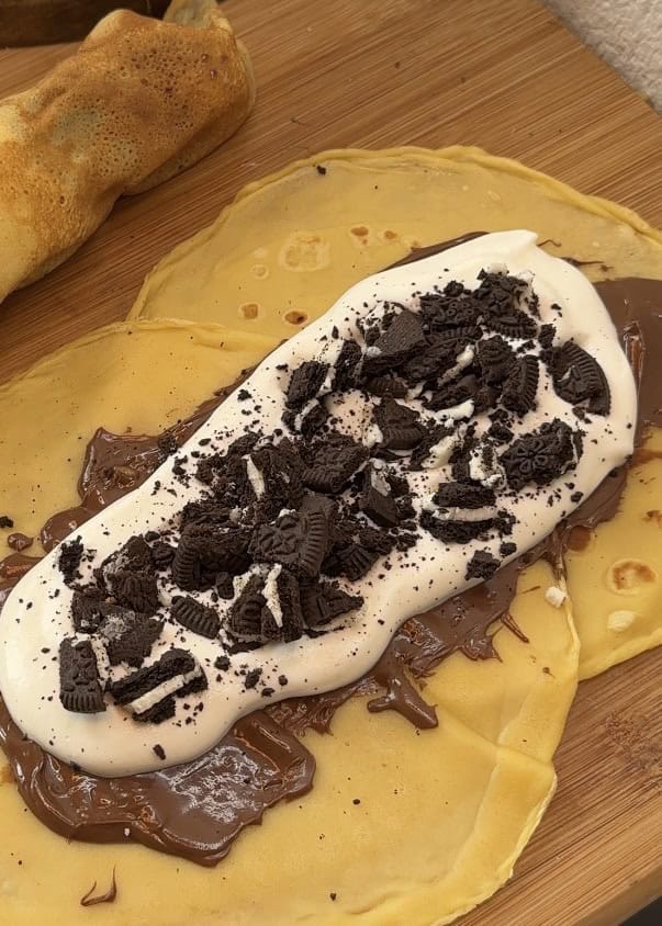 Chocolate crepe filled with whipped cream and crushed Oreo cookies, ready to be rolled, presented on a rustic wooden table