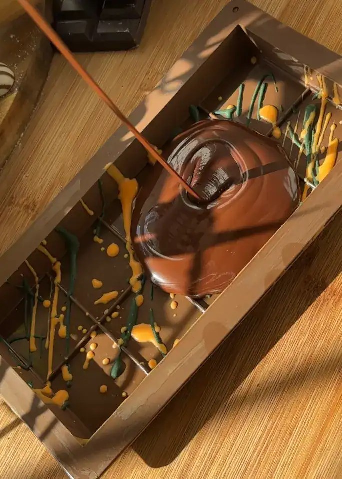 Pouring rich, melted chocolate over a mold splashed with dark green and orange-colored white chocolate to create homemade Dubai chocolate bars.