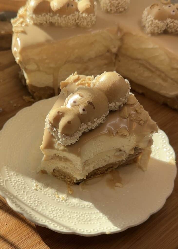 A slice of Happy Hippo No-Bake Cheesecake on a plate, featuring a crunchy biscuit base, creamy hazelnut cheesecake filling, Kinder-filled interior, and topped with Kinder Bueno spread, wafer rolls, Happy Hippo, and pearl sugar