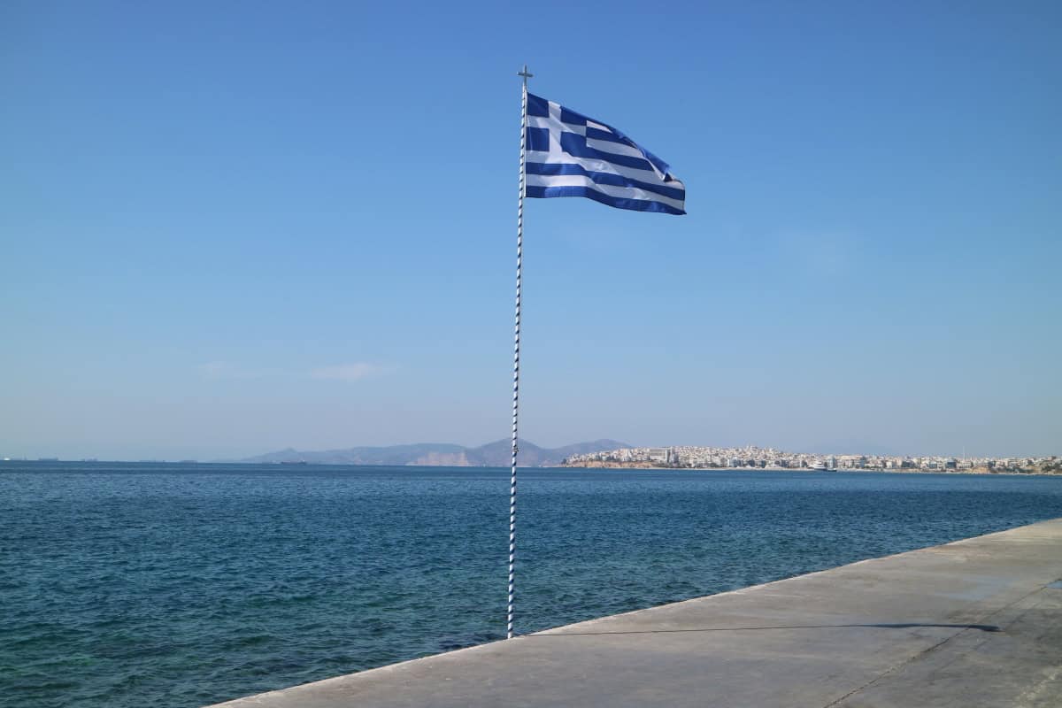 Vibrant Greek flag waving over coastal waters, showcasing Greek culture and scenic views from The Wandering Woodward.