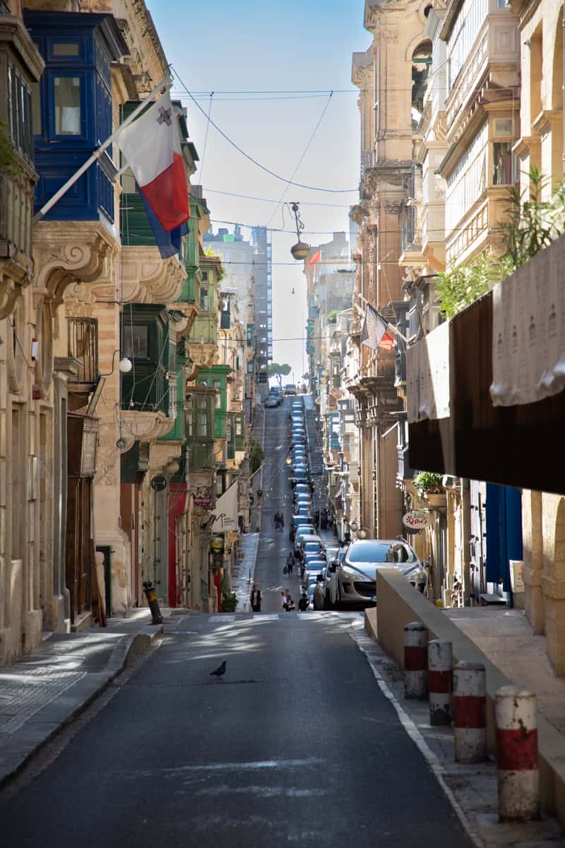 Old hillside street in Valletta, Malta with historic buildings and parked cars.