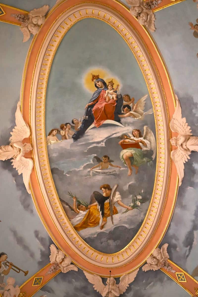 Angel and religious fresco painting on ceiling with ornate pink and gold frame at The Wandering Woodward, showcasing baroque style artwork.