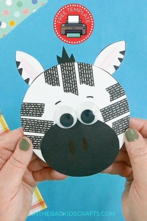 A person holding a completed preschool zebra craft, a fun fine motor activity featuring a round paper zebra face with black stripes, googly eyes, and a blue background.