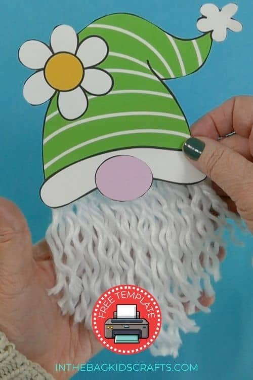 A pair of hands holding a completed gnome craft, featuring a green striped hat with a flower and a textured yarn beard, making it a fun and easy craft for kids.