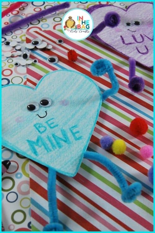A colorful Valentine craft featuring a heart craft made with googly eyes, pipe cleaners, and pom-poms on a striped background, showcasing an easy craft for kids and a fun paper plate craft idea.