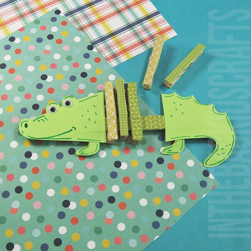 A green paper alligator made with painted clothespins and cardstock is displayed on colorful patterned backgrounds, showing a playful and creative alligator craft for kids.