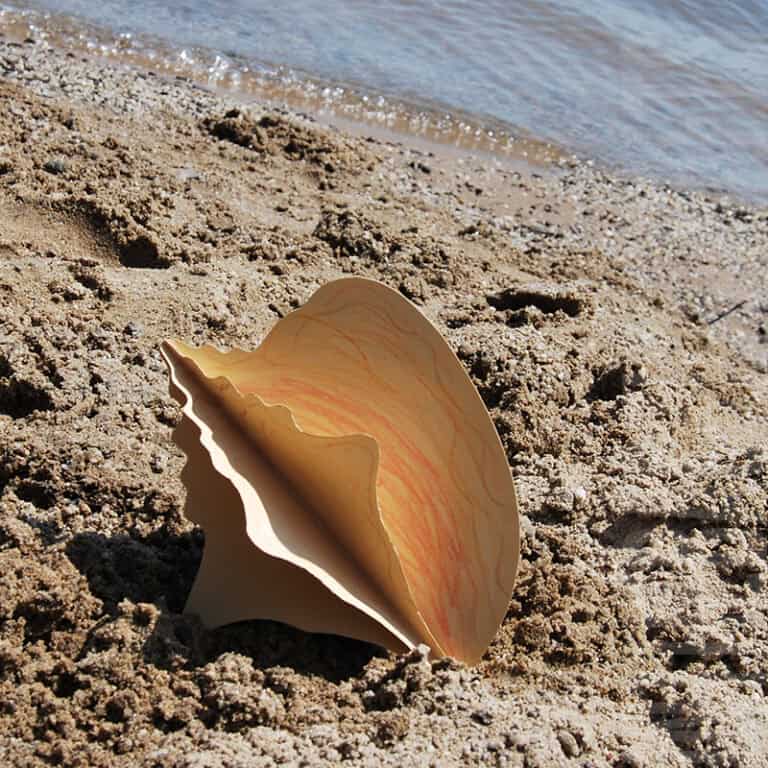 Two hands holding a three-dimensional paper conch shell colored in warm tones, representing a creative conch shell craft and beach craft for kids.