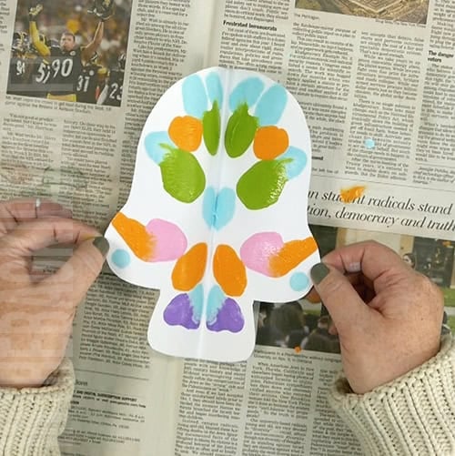 An image showing a colorful paper skull craft for kids, perfect as a Day of the Dead craft activity.  step 3 opening the squish paint art 
