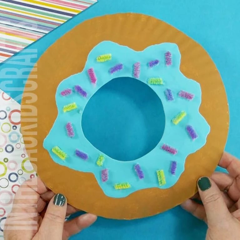 A colorful donut craft for kids made from a paper plate craft, decorated with blue "frosting" and pipe cleaner sprinkles, creating a fun and easy food craft activity.