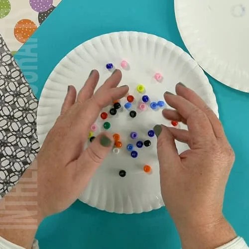 An image showing a simple paper plate ghost craft, perfect for an easy Halloween craft and fun preschool activity for kids. step 2 adding beads to the center 