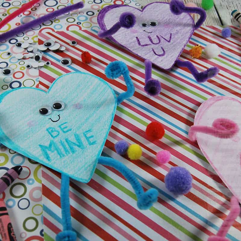 A colorful Valentine craft featuring a heart craft made with googly eyes, pipe cleaners, and pom-poms on a striped background, showcasing an easy craft for kids and a fun paper plate craft idea.