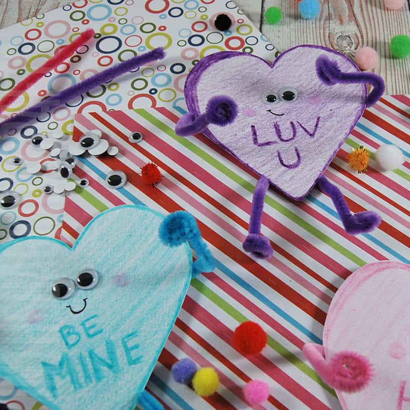A colorful Valentine craft featuring a heart craft made with googly eyes, pipe cleaners, and pom-poms on a striped background, showcasing an easy craft for kids and a fun paper plate craft idea.