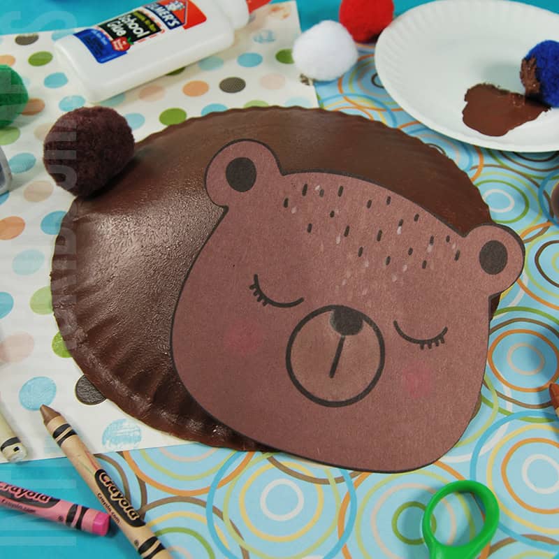 An adorable bear craft for kids made from a painted paper plate, featuring a hibernating animal design, surrounded by craft supplies like crayons, glue, and pom-poms.