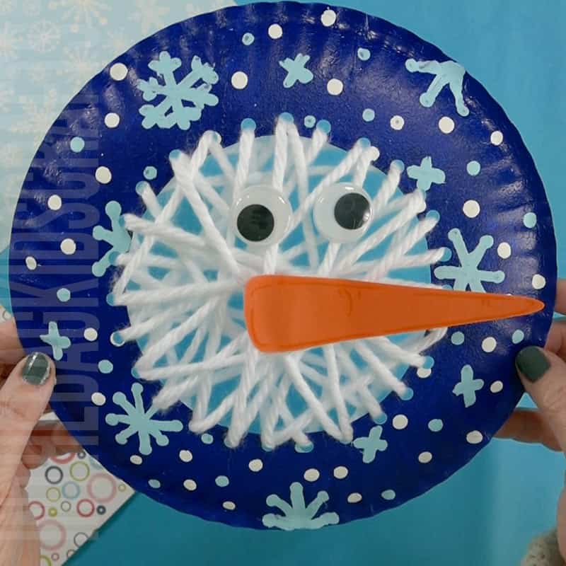 A colorful yarn snowman craft for kids featuring a blue paper plate decorated with white yarn lacing, snowflakes, googly eyes, and an orange carrot nose, perfect as a fun winter craft activity.