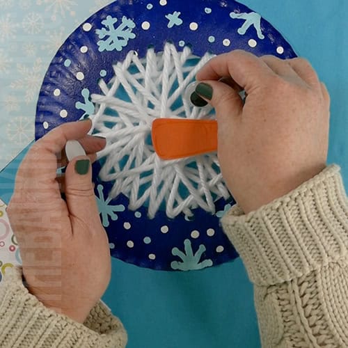 A colorful yarn snowman craft for kids featuring a blue paper plate decorated with white yarn lacing, snowflakes, googly eyes, and an orange carrot nose, perfect as a fun winter craft activity.