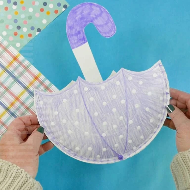 Child holding a handmade purple paper plate umbrella craft with polka dots, designed as a fun Spring craft and pouch project.